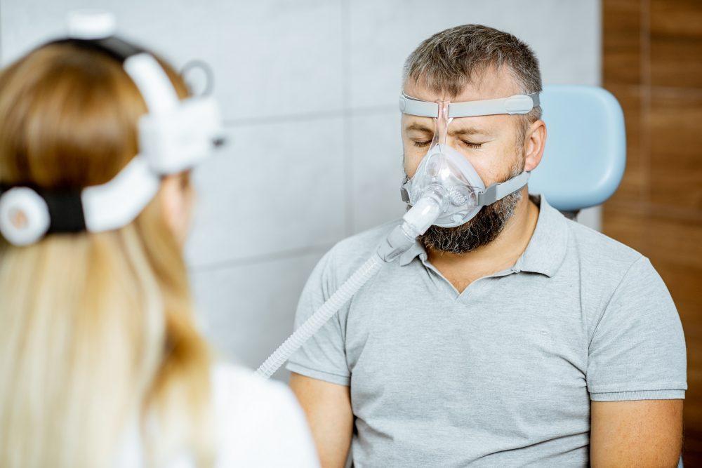Mies lääkärin tai hoitajan vastaanotolla. Mies testaa uniapnean hoitoon tarkoitetun hengityslaitteen maskia.
