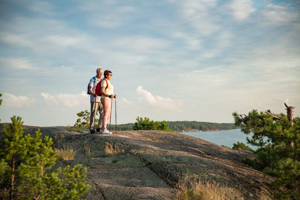 Mies ja nainen sauvakävelyllä. Pari on kiivennyt korkean kallion päälle, josta he ihailevat kaunista järvimaisemaa.