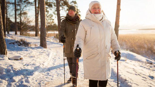 Kaksi henkilöä sauvakävelylenkillä. Kuvan henkilöt kävelevät pitkin lumista metsäpolkua keskellä talvea.