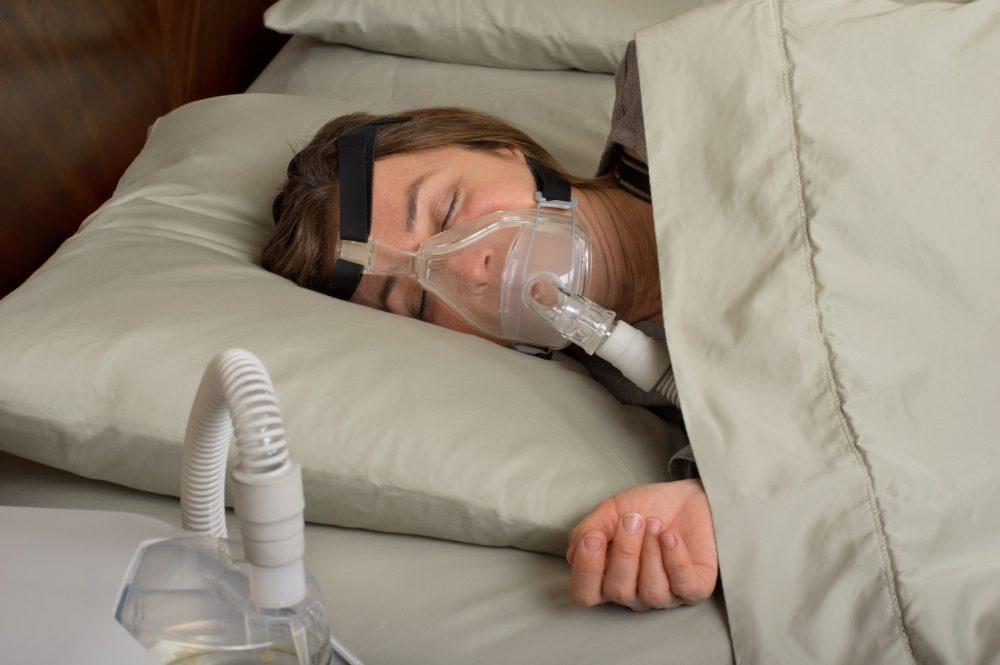 Nainen nukkuu CPAP-laitteen nenä-suumaski kasvoillaan.