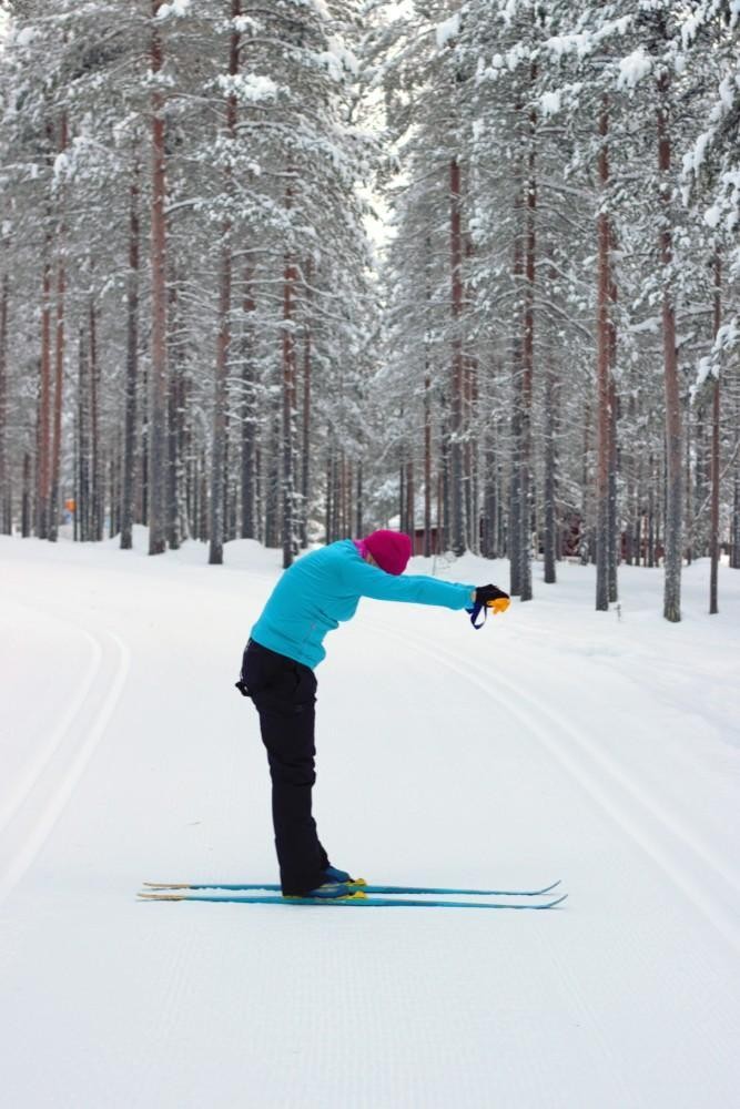 Nainen suksilla nojaa  ja pyöristää yläselkäänsä sauvat edessään.