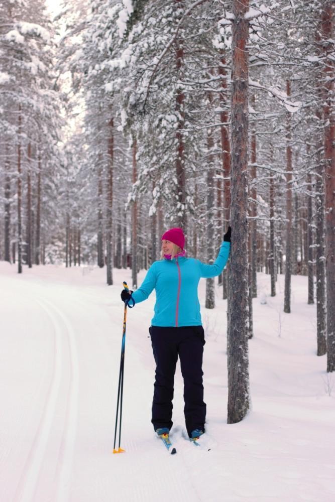 Nainen suksilla. Toinen käsi nojaa puuhun niin, että rintalihas venyy.